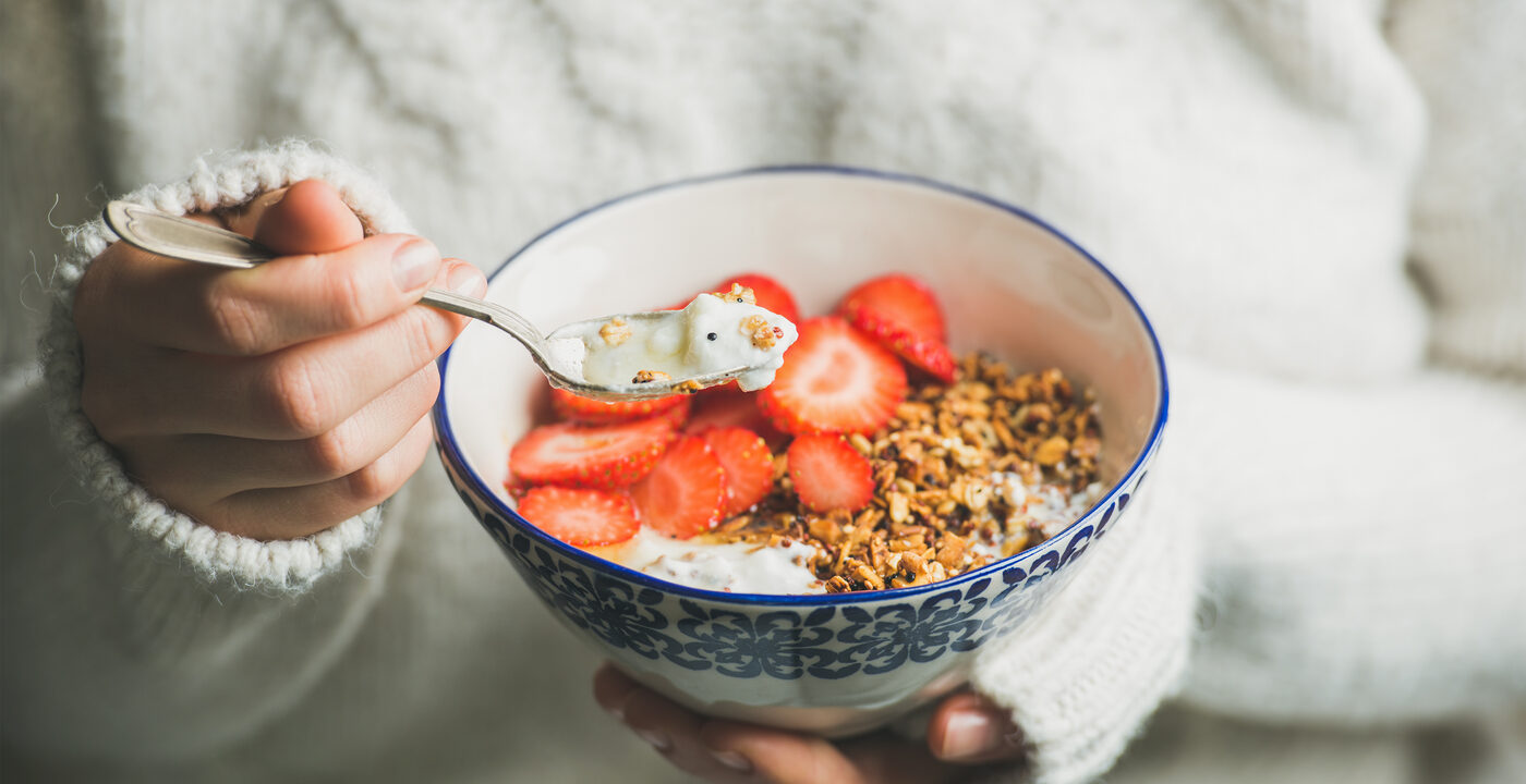 Healthy breakfast greek yogurt, granola and strawberry bowl in hands of woman wearing white loose knitted woolen sweater, selective focus. Clean eating, healthy, vegetarian, dieting food concept
