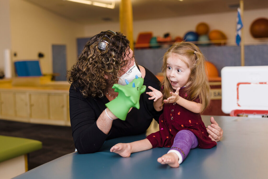 Toddler in a red dress and purple orthotics on her left leg reacts with a surprised and happy look as her physical therapist holds up a triceratops hand puppet