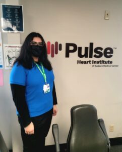 Woman with dark hair and face mask stands in front of Pulse Heart Institute of Auburn Medical Center wall sign