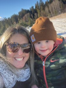 Mom in sunglasses and toddler in beanie smile at the camera in a sunny woodsy area