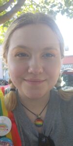 Portrait of a person with blonde braids and a rainbow Pride necklace and pins