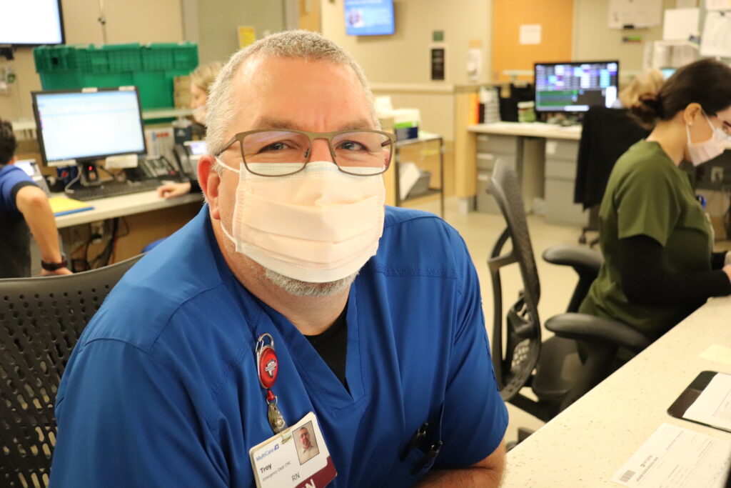 Emergency department nurse wearing face mask and glasses sits at a nurse station