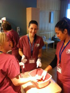 Students wearing nurse scrubs and gloves surround a table with a pink tub, laughing