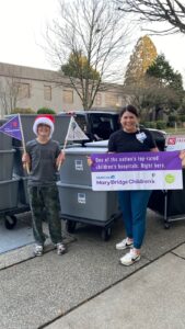 Boy in red plaid pullover smiles in front of a car trunk loaded with boxes of toys