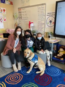 Social worker, child life specialist, child patient and golden retriever dog with blue vest pose for a picture in a classroom