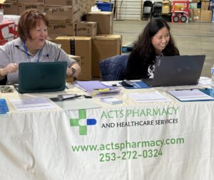 Two staff members sit behind a table with laptops and forms at an “ACTS Pharmacy and Healthcare Services” booth inside a warehouse with stacked supply boxes.