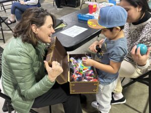 Adult seated at a table opens a small chest filled with toys while a child chooses an item; another adult stands nearby in a community event setting.