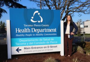Person stands next to a large outdoor sign reading “Tacoma-Pierce County Health Department” with trees and buildings in the background.