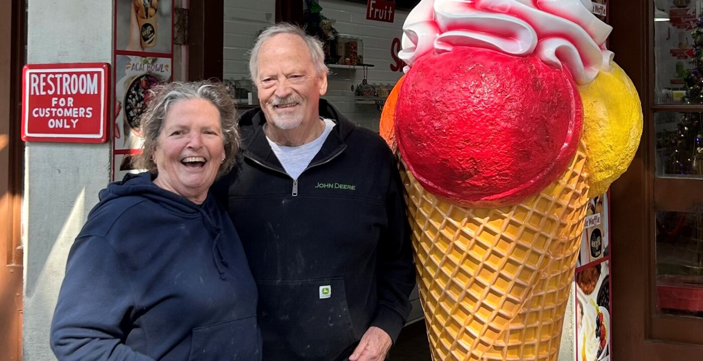 Two people stand outside a storefront next to a large decorative ice cream cone statue with pink and red scoops, holding glasses and wearing casual clothing.