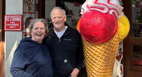 Two people stand outside a storefront next to a large decorative ice cream cone statue with pink and red scoops, holding glasses and wearing casual clothing.