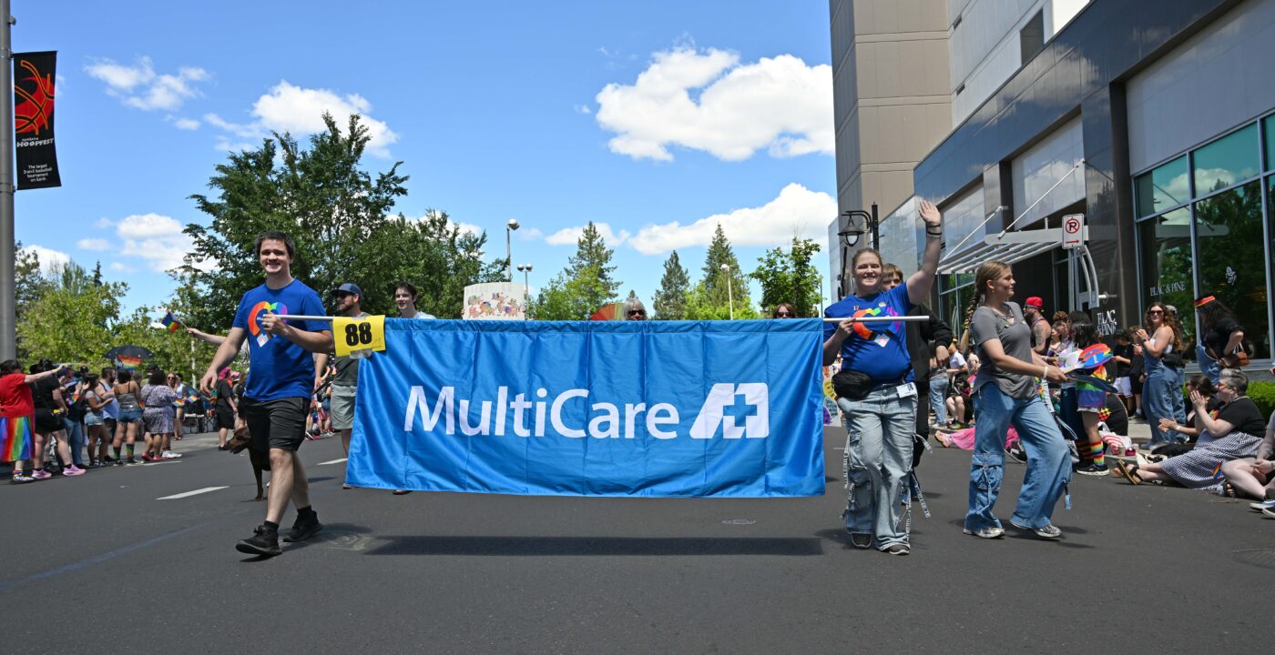 Parade participants carry a large blue banner reading “MultiCare” while walking down a street during a Pride event, with crowds watching on both sides.