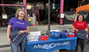 Two people stand behind a table covered with a blue “MultiCare” tablecloth and promotional items, holding branded tote bags at an outdoor event.