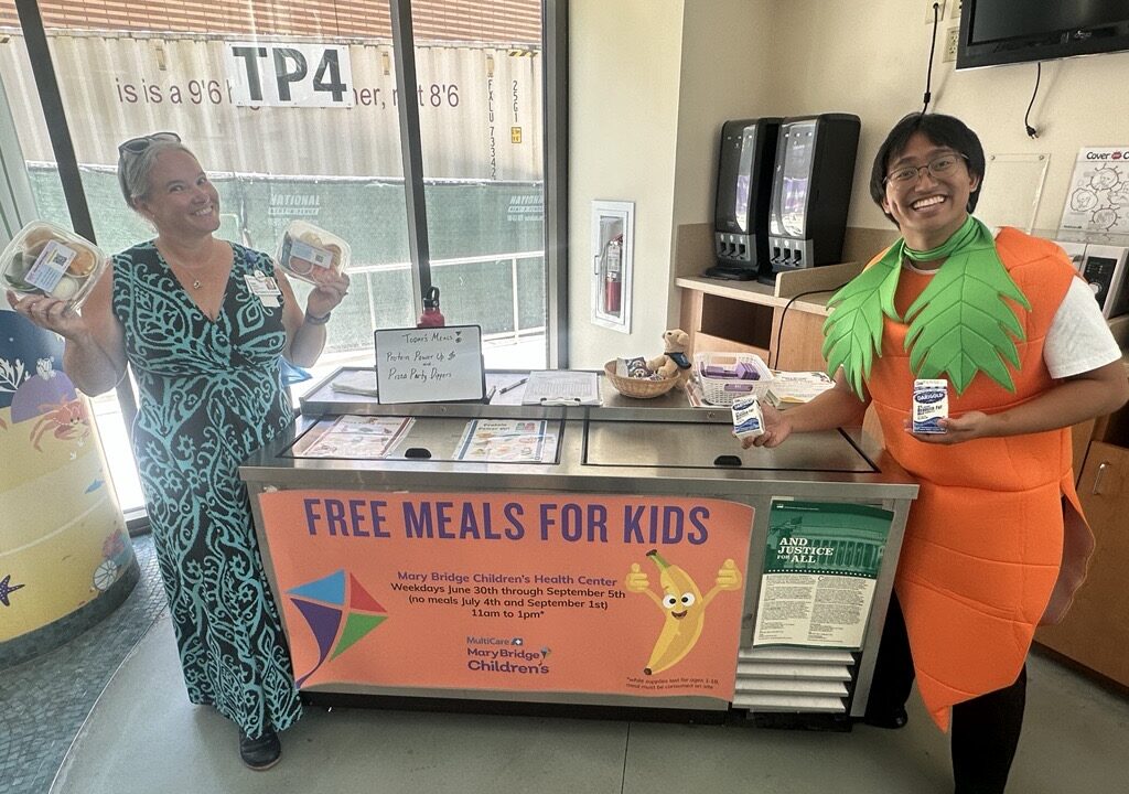 Two adults standing at a “Free Meals for Kids” distribution table indoors, each holding packaged food items, with signage indicating a summer meal program at Mary Bridge Children’s Health Center.