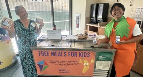 Two adults standing at a “Free Meals for Kids” distribution table indoors, each holding packaged food items, with signage indicating a summer meal program at Mary Bridge Children’s Health Center.