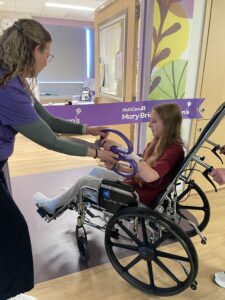 A child seated in a wheelchair holds purple ribbon scissors while another person assists, with a “MultiCare Mary Bridge Children’s” ribbon stretched across a hospital room doorway.