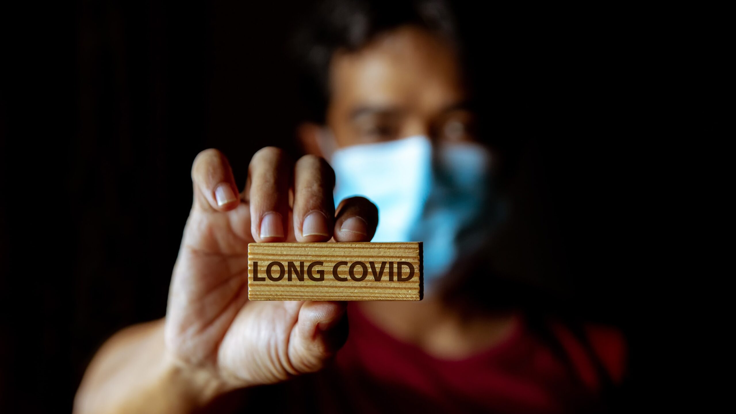 A masked person holding up a block that says long covid on it.